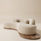Sofá curvo orgánico Ámbar tapizado en tela texturizada beige claro con base de madera natural extendida y dos cojines esféricos.