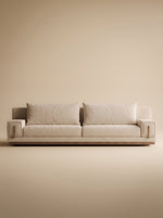 Vista frontal de sofá contemporáneo modelo Dominic tapizado en tejido texturizado tono beige arena, con dos cojines amplios de respaldo, apoyabrazos rectilíneos con detalle de hebilla o herraje vertical en acabado dorado cepillado y base zócalo de madera natural, ambientado sobre fondo neutro de estudio.