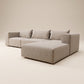 Vista frontal de sofá modular Magnum con configuración de chaise longue a la derecha, tapizado en tejido texturizado tono crema claro, con tres cojines de respaldo sueltos, ribete decorativo en color marrón oscuro en los contornos y patas cortas de madera natural, presentado en un estudio minimalista.
