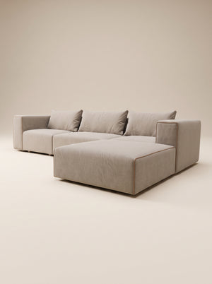 Vista frontal de sofá modular Magnum con configuración de chaise longue a la derecha, tapizado en tejido texturizado tono crema claro, con tres cojines de respaldo sueltos, ribete decorativo en color marrón oscuro en los contornos y patas cortas de madera natural, presentado en un estudio minimalista.