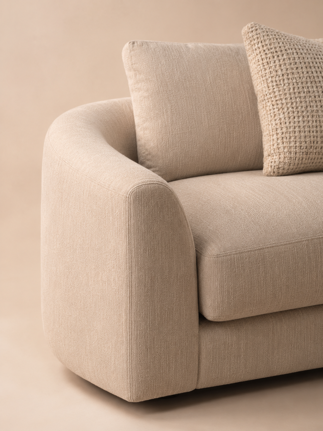 Sofá curvo de dos plazas Oasis tapizado en tela beige texturizada con cojines decorativos a juego, un cojín circular oscuro y placa dorada con la marca.
