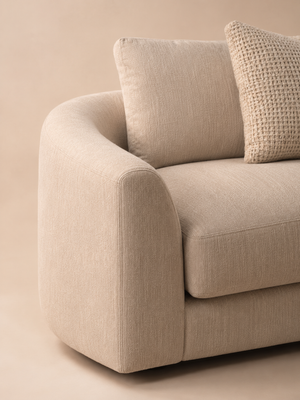 Sofá curvo de dos plazas Oasis tapizado en tela beige texturizada con cojines decorativos a juego, un cojín circular oscuro y placa dorada con la marca.