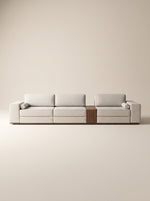 Sofá modular Reggio tapizado en tela neutra texturizada con mesa lateral integrada de madera natural y cojines cilíndricos sobre fondo blanco.