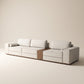 Sofá modular Reggio tapizado en tela neutra texturizada con mesa lateral integrada de madera natural y cojines cilíndricos sobre fondo blanco.